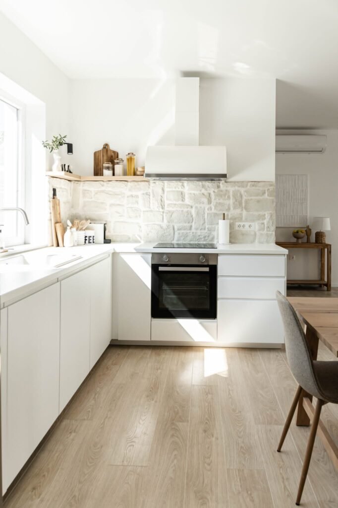 A modern kitchen with white cabinets, wooden flooring, and a stone backsplash. includes a sink, oven, and chimney with natural light streaming in.