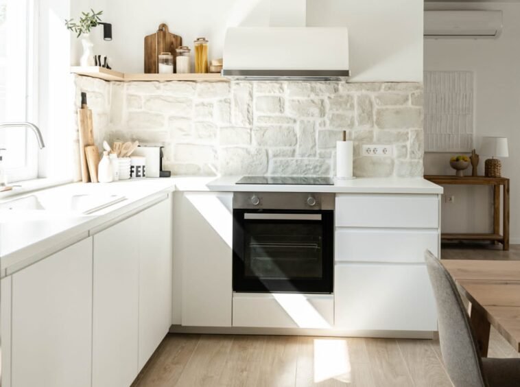 A modern kitchen with white cabinets, wooden flooring, and a stone backsplash. includes a sink, oven, and chimney with natural light streaming in.