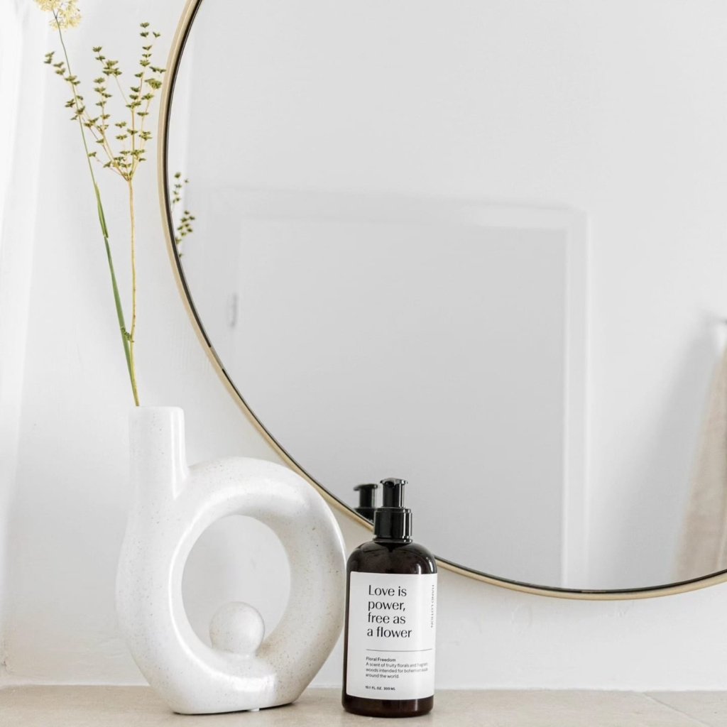 A minimalist interior scene with a decorative round mirror, a white sculptural vase with dried flowers, and a bottle labeled "love is power, free as a flower" on a pale surface against a white wall.