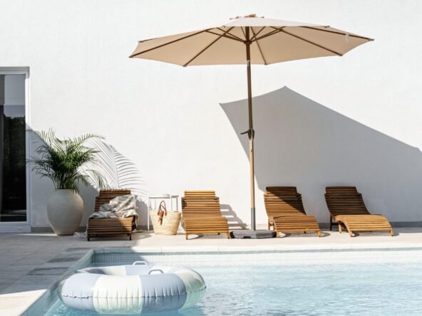 A serene poolside setting with four wooden loungers under a large beige umbrella, a float in the water, and a potted plant beside a white building on a sunny day.