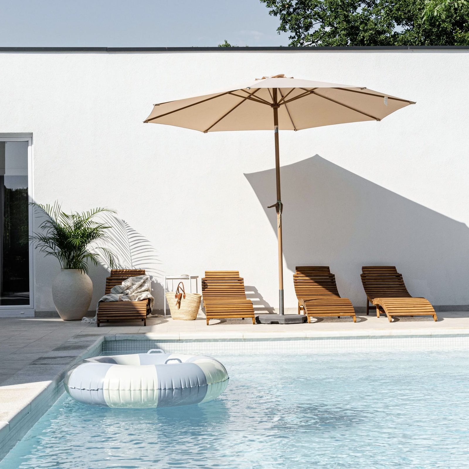 A serene poolside setting with four wooden loungers under a large beige umbrella, a float in the water, and a potted plant beside a white building on a sunny day.