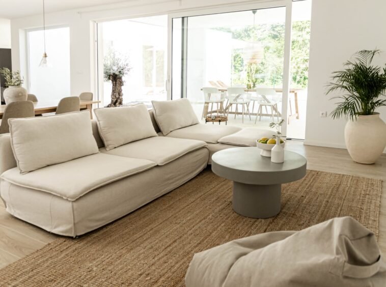 Spacious, modern living room with a large beige sofa, gray coffee table, potted plants, and an open dining area in the background. natural light fills the room through large glass doors.