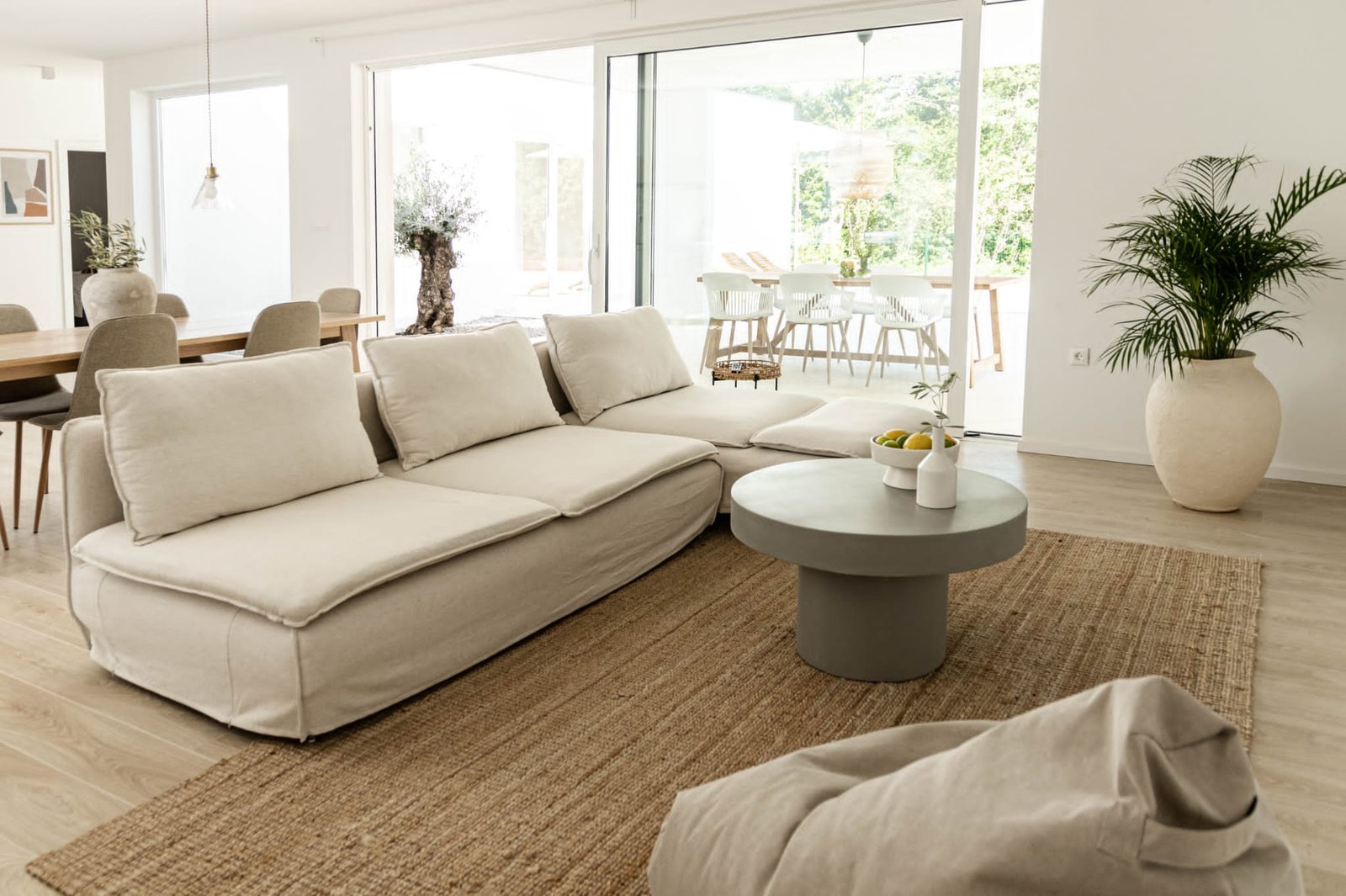 Spacious, modern living room with a large beige sofa, gray coffee table, potted plants, and an open dining area in the background. natural light fills the room through large glass doors.