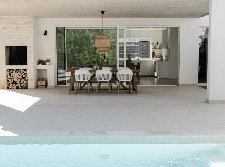 Modern outdoor dining area by a pool, featuring a stylish table with chairs and an open view of a kitchen inside a house with stone and white walls.