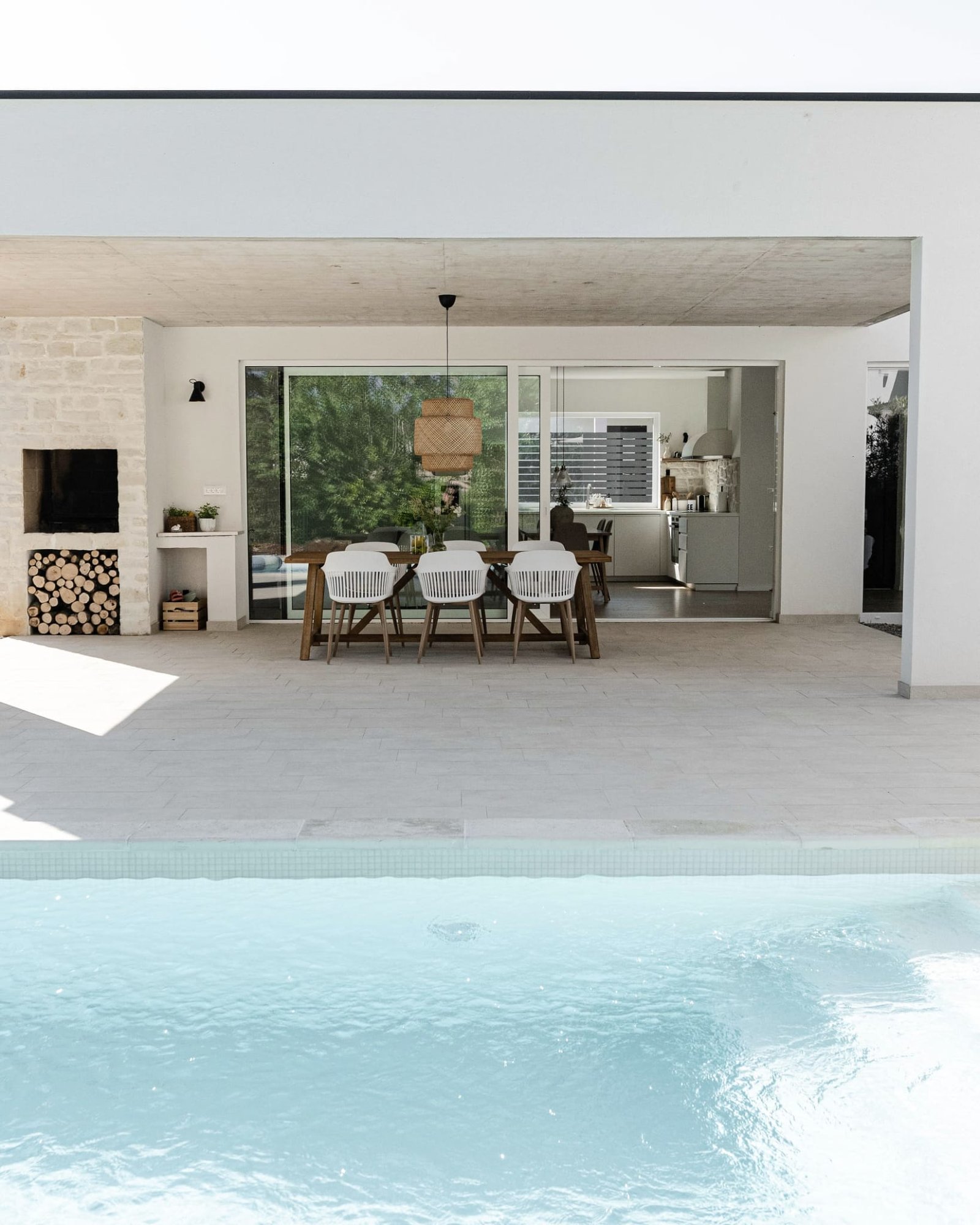 Modern outdoor dining area by a pool, featuring a stylish table with chairs and an open view of a kitchen inside a house with stone and white walls.