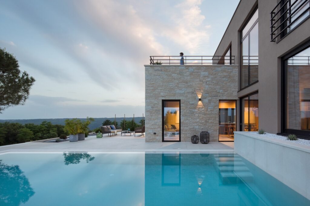 Modern two-story house in Istria with large windows and stone facade at twilight, featuring an infinity pool in the foreground and a serene natural backdrop.