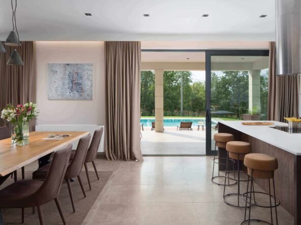 A spacious modern kitchen and dining area with large glass doors opening to a poolside view, featuring a sleek breakfast bar, wooden dining table, and elegant decor.