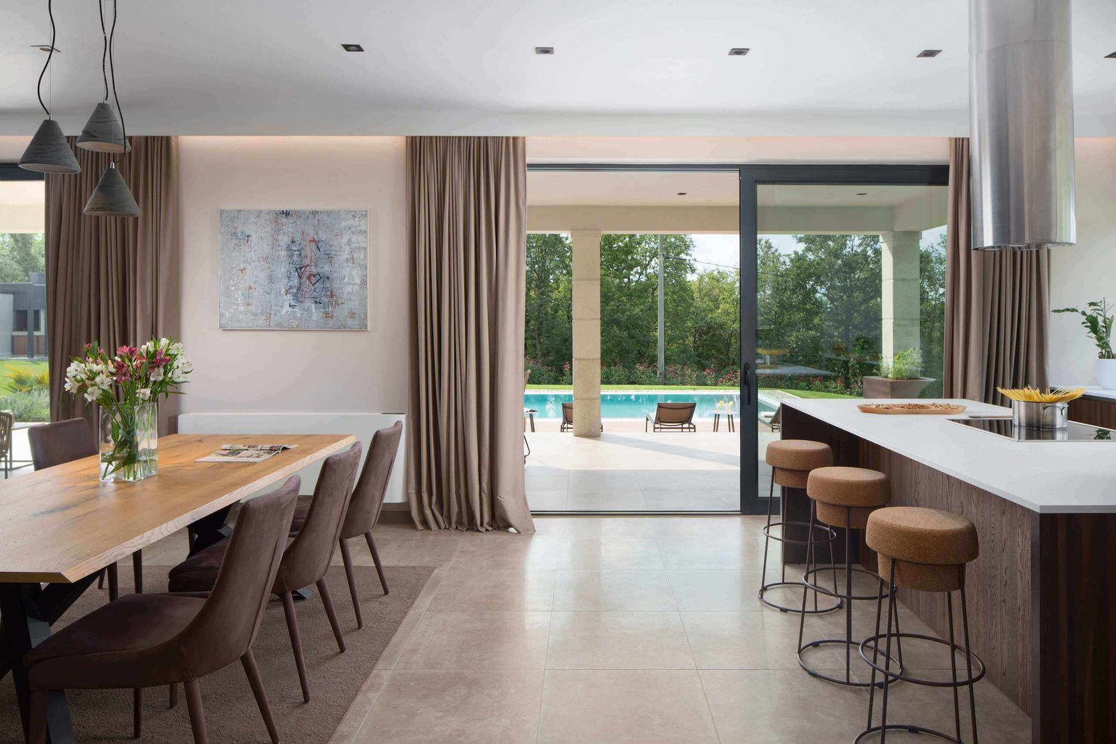 A spacious modern kitchen and dining area with large glass doors opening to a poolside view, featuring a sleek breakfast bar, wooden dining table, and elegant decor.