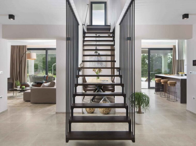 Modern interior of a spacious home with sleek metal stairs leading to the upper level, large windows, and an open-plan living area with minimalist decor.