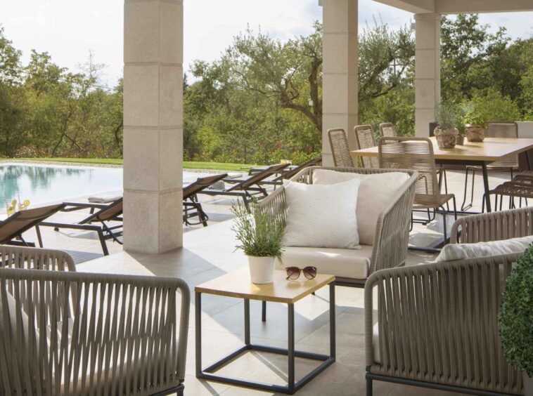 A luxurious poolside patio with elegant outdoor furniture including loungers, chairs, and a dining area, surrounded by lush greenery in a serene setting.