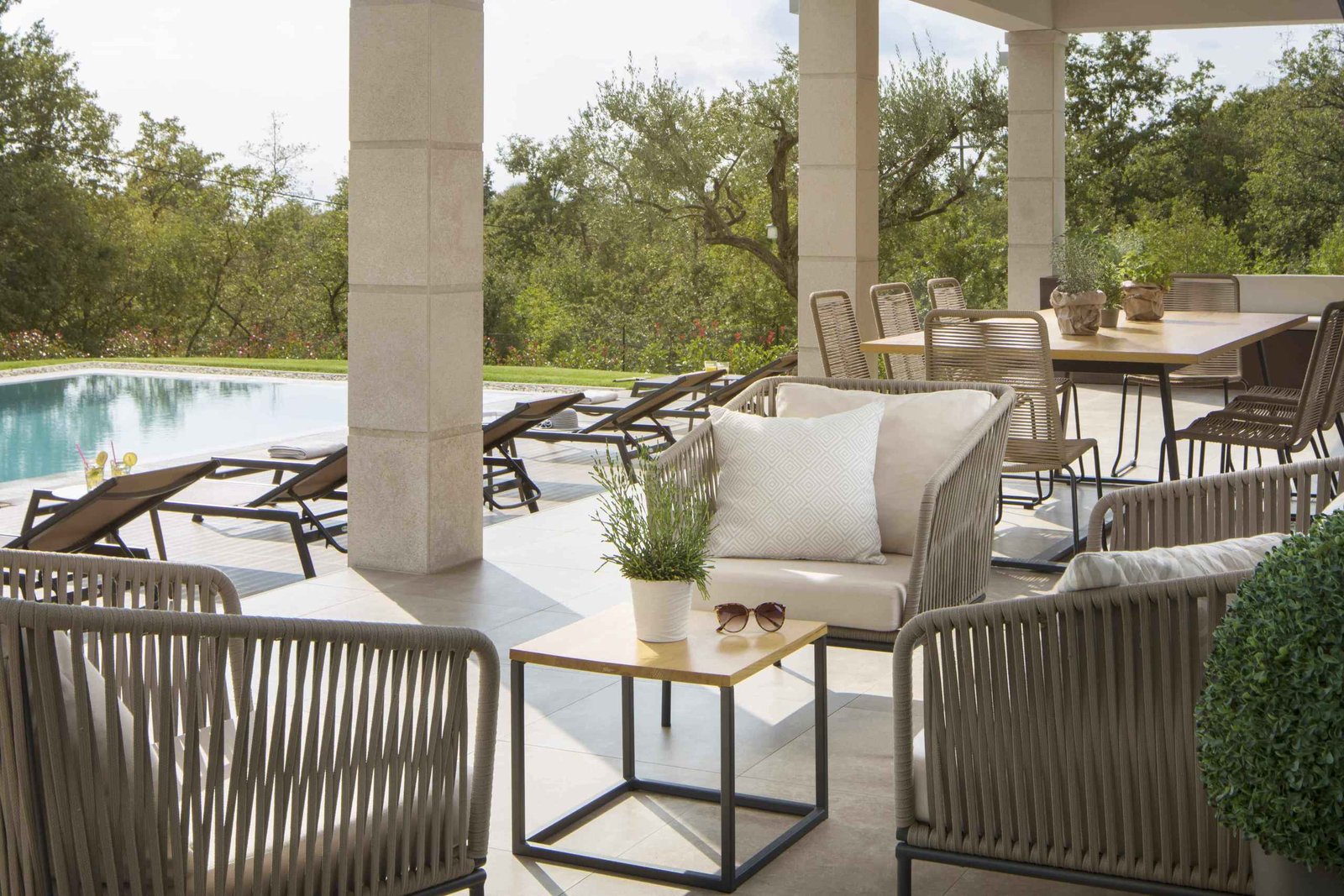 A luxurious poolside patio with elegant outdoor furniture including loungers, chairs, and a dining area, surrounded by lush greenery in a serene setting.