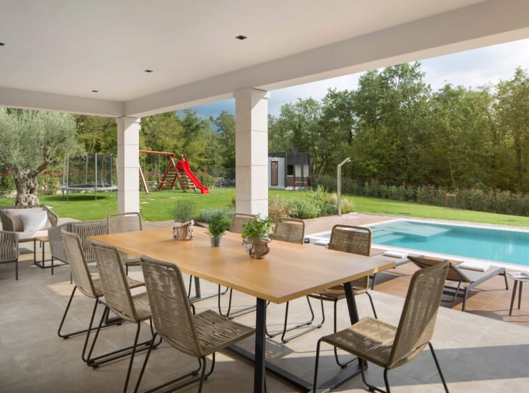 An elegant outdoor dining area with a large wooden table and wicker chairs, overlooking a swimming pool and lush garden with a red slide in the background, all under a covered patio.