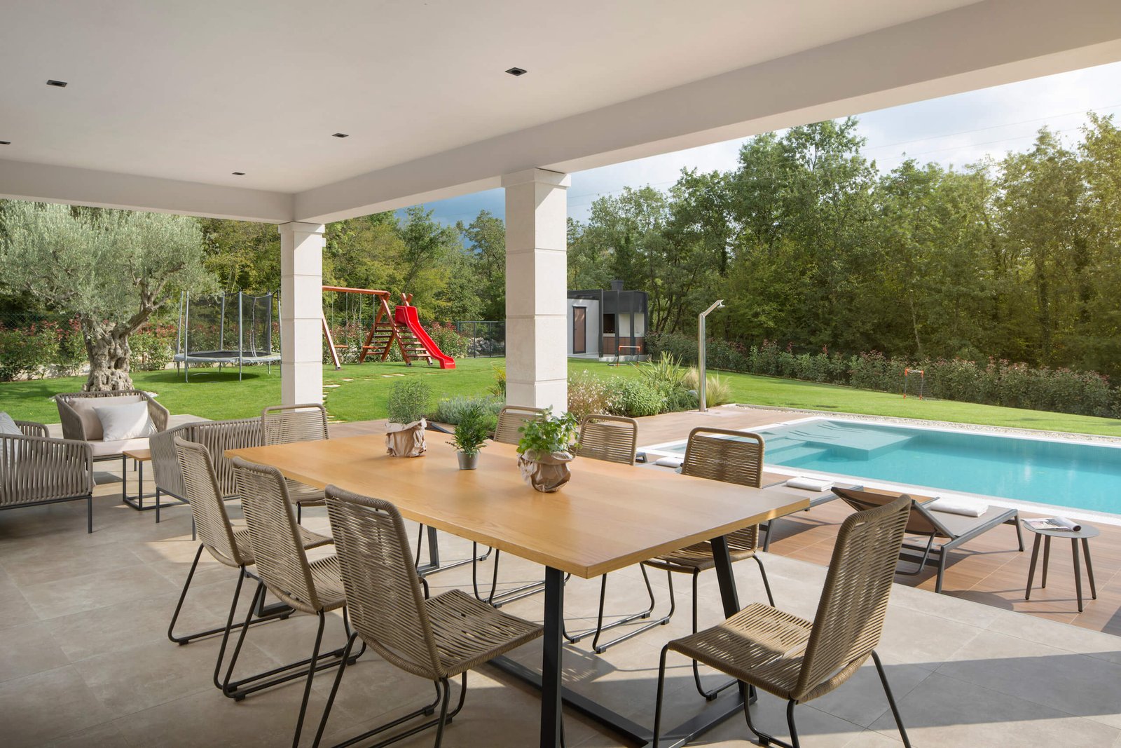 An elegant outdoor dining area with a large wooden table and wicker chairs, overlooking a swimming pool and lush garden with a red slide in the background, all under a covered patio.