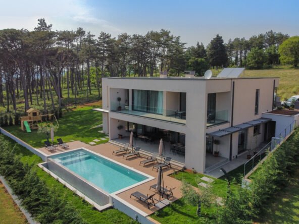 Aerial view of a modern two-story house with a large swimming pool, surrounded by lush greenery. the house features large glass windows, a balcony, and a well-maintained lawn with patio furniture.